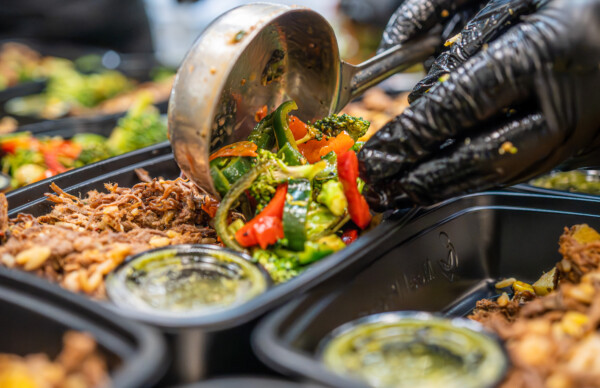 Chef ladling cooked broccoli, peppers, and zucchini into a Meal Prep Sunday premade meal with shredded beef and rice